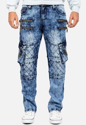 Blaue Denim-Cargohosen mit gesteppter Patches, mehreren Reißverschlusstaschen und mit Knöpfen verschlossenen Klappentaschen an den Seiten, getragen mit weißen Sneakers.