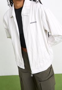 Bílá bunda na zip s vertikálními tmavě modrými pruhy, krátkými rukávy a plochým límcem, s malým černým logem adidas na levé straně hrudi.