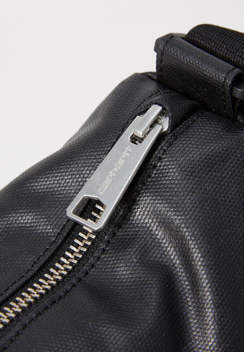 Sac en tissu texturé noir avec un détail en zip métal argenté, doté d'un tirette hexagonale et d'un attachement de bandoulière tissée.
