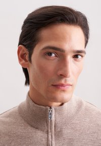 Junger Mann mit dunklem Haar, der einen beigen Reißverschluss-Pullover trägt und direkt voraus blickt, vor einem schlichten hellen Hintergrund.