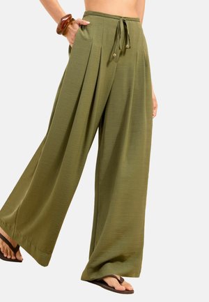 weite olivgrüne Hose mit vorderen Falten und einem Tunnelzugbund, getragen mit dunklen Flip-Flops und einem braunen Armband am Handgelenk.