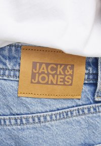 Hnedá kožená nášivka na džínsoch s textom "JACK & JONES" prišitá na vyblednutý modrý denimový pás pod bielou košeľou.