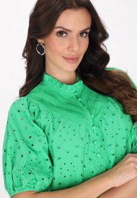 Grüne bestickte Bluse mit Puffärmeln, Knopfleiste und rüschigem Kragen. Der Stoff hat Spitzen-Details und eine strukturierte Oberfläche.