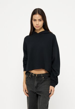 Junge Frau mit langen dunklen Haaren, trägt Creolen, einen schwarzen, kurz geschnittenen Hoodie und eine hochtaillierte dunkle Jeans mit schwarzem Gürtel, steht vor neutralem Hintergrund.