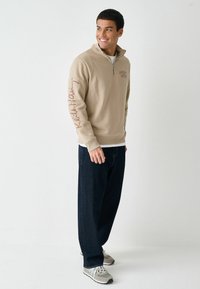 Beige lynlås-sweatshirt med høj krave, med et logo på brystet og "Keith Haring" tekst i brun på ærmet. Mørke denimbukser og grå sneakers.
