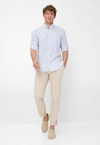 Camicia a righe blu chiaro e bianca con maniche arrotolate, abbinata a pantaloni beige e scarpe slip-on, su uno sfondo neutro.