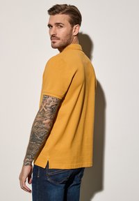 Musztardowa żółta koszulka polo z krótkimi rękawami, bocznymi rozcięciami i klasycznym kołnierzykiem, w połączeniu z ciemnoniebieskimi dżinsami. Prezentuje widoczne tatuaże.