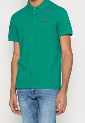 Mężczyzna w zielonej koszulce polo z krótkim rękawem, trzema guzikami oraz małym czerwonym logo Levi's, w połączeniu z jasnoniebieskimi dżinsami.