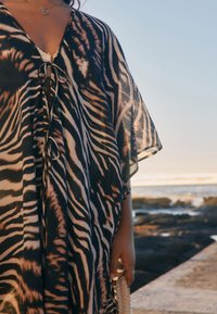 Durchsichtiger Kaftan mit schwarzem und beigem Zebra-Print, V-Ausschnitt und Bindebändern vorne. Weiche Textur, fließendes Design und lockere Passform.
