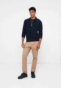 Pulover tricotat bleumarin cu guler cu fermoar, asociat cu pantaloni tapered bej deschis și adidași negri, pe un fundal alb simplu.
