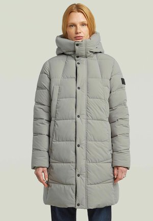 Vrouw draagt een lichtgrijze doorgestikte lange donsjacket met hoge kraag en drukknopen, staand tegen een effen lichte achtergrond.