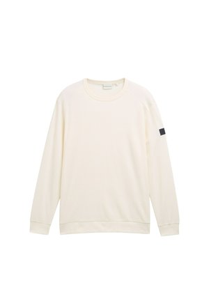 Felpa a maniche lunghe color crema, texture a coste, scollo rotondo, maniche raglan, presenta una piccola toppa nera sulla manica sinistra.