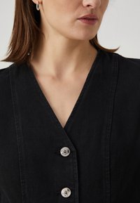 Μαύρο τζιν φόρεμα με V-ντεκολτέ και δύο μεταλλικά κουμπιά snap. Το ύφασμα έχει λεία υφή και διαθέτει εφαρμοστή σιλουέτα.