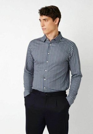 Mann trägt ein blau-weiß vertikal gestreiftes Hemd, das in schwarze Hose gesteckt ist, Hände in den Taschen, schaut zur Seite.