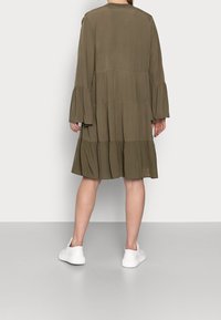 Olivgrön, lager-på-lager, midikjol med utställda ärmar, tillverkad av lättviktsmaterial. Bärs med vita sneakers. Minimalistisk design.