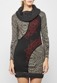 Schwarzes, gemustertes Kleid mit langen Ärmeln, Cowl-Neck und roter Stickerei. Es zeichnet sich durch komplexe geometrische und Paisley-Muster aus und hat eine figurbetonte Form.