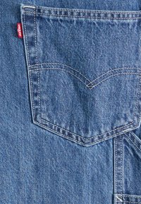 Τσέπη πίσω από μπλε τζιν Levi's με χαρακτηριστικό ραμμένο σχέδιο, που διαθέτει κόκκινη ετικέτα επωνυμίας και υφασμάτινη υφή.