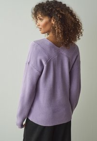 Maglione lilla a coste con scollatura a V e cuciture delle spalle testurizzate, caratterizzato da un fit rilassato e maniche lunghe. Pantaloni neri a righe sottili in vista sotto.