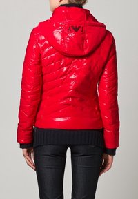 Röd blank puffjacka med huva, kviltad design och svart ribbad mudd. Logotyp på ryggen. Stylad med mörka jeans.