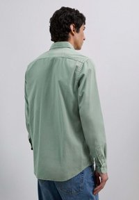 Hombre vistiendo una camisa de manga larga verde claro y jeans azules, de pie de espaldas contra un fondo gris liso.