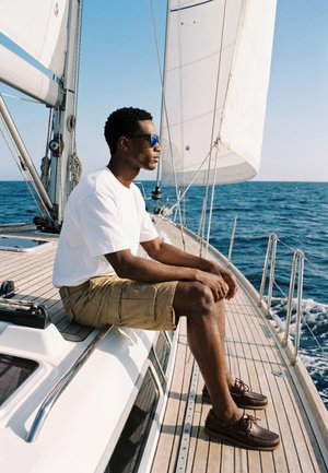Un homme portant des lunettes de soleil et un short est assis au bord du pont d'un voilier, regardant la mer sous un ciel bleu clair.