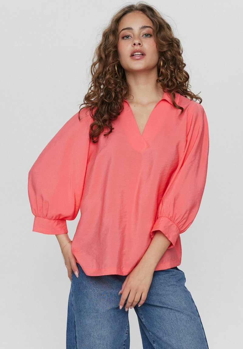 Blusa color corallo con scollatura a V, maniche lunghe a sbuffo e vestibilità rilassata. Tessuto liscio con un minimo di lucentezza. Abbinata a pantaloni in denim.