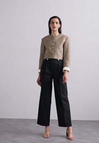 Getextureerde bruine cropped jacket met gouden knopen, witte manchetten; gecombineerd met hoge zwarte broeken en strappy gouden sandalen.