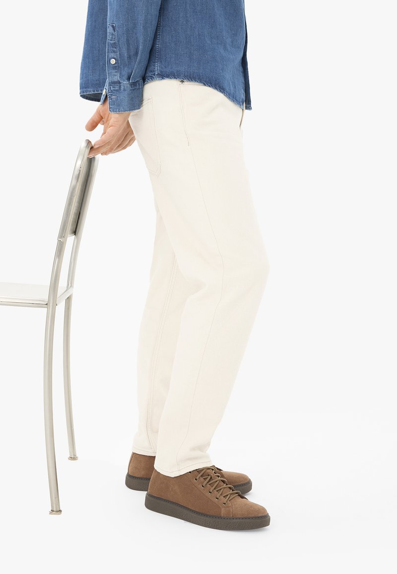 Personne portant un pantalon blanc et des chaussures marron, tenant une chaise en métal par le dossier, debout sur un fond blanc.