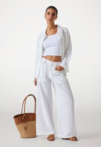 Vit kortärmad topp, vita lösa byxor med fickor, vit oversized jacka och en brun och halmvävds toteväska. Bruna flata sandaler.