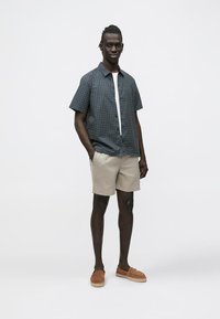 Navyblauw shirt met korte mouwen en geometrisch patroon, lichtgekleurde korte broek en tan-kleurige instapschoenen. Model staat tegen een effen witte achtergrond.