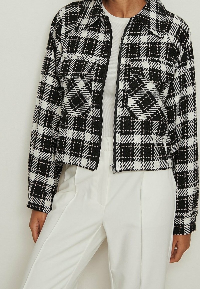 Veste courte à carreaux noir et blanc avec fermeture éclair, deux poches poitrine, et tissu texturé contrastant. Portée sur un haut blanc.