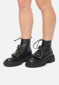 Zwarte leren veterlaarzen tot de enkel met grote metalen kettingaccenten, ronde neus en stevige rubberen zool. Zijrits voor gemakkelijk aan- en uittrekken.