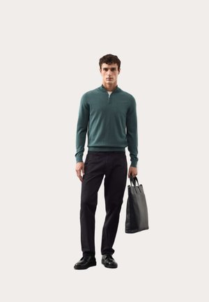 Man draagt een tealkleurige trui met een rits tot een kwart, zwarte broek en zwarte schoenen, en houdt een zwarte leren handtas vast tegen een effen achtergrond.