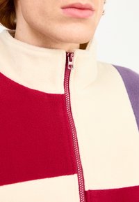 Farvet blokket fleecejakke med nuancer af burgundy, creme og lavendel. Høj krave med en kontrastfarvet lynlåsdetalje. Blød tekstur.