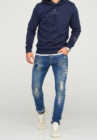 Mörkblå hoodie med snörning, med en logga fram. Bärs med slitna blå jeans som har hål. Avslutas med beiga sneakers.