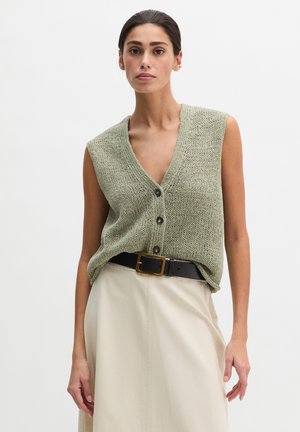 Kobieta w zielonej, bezrękawnikowej dzianinowej kamizelce zapinanej na guziki, wpuszczonej w jasnobrązową spódnicę z czarnym paskiem, stojąca na jasnym, jednolitym tle.