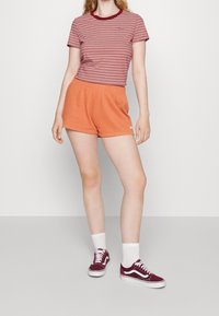 Osoba ubrana w czerwono-biały pasiasty T-shirt, pomarańczowe szorty, białe skarpetki i bordowe trampki, stojąca na tle białej ściany.