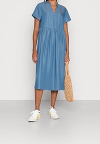 Džínová midi šaty světle modré barvy, s krátkými rukávy, výstřihem do V a nařaseným pasem. Spárovány s bílými teniskami a tkanou kabelkou.
