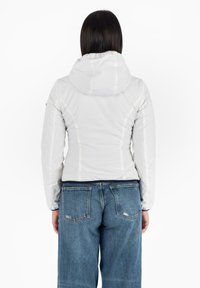 Witte, geïsoleerde jas met een capuchon, met zichtbare stikdetails, gedragen over een blauwe spijkerbroek. De stof lijkt glad en lichtgewicht te zijn.