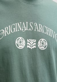 Μαλακό πράσινο μπλουζάκι με λευκό κείμενο "ORIGINALS ARCHIV" και τρία λευκά λουλουδένια σύμβολα κάτω από το κείμενο.