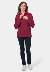 Magenta gestrickter Pullover mit hohem Kragen und strukturiertem Muster, kombiniert mit dunkelblauen Skinny-Jeans und weißen Sneakers.