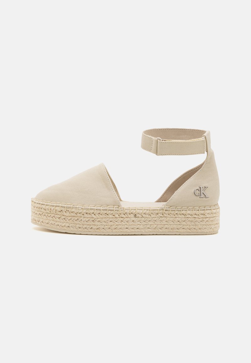 Espadrille beige avec une tige en toile, une bride à la cheville et une semelle en jute tressé. Elle présente un bout arrondi et un détail de logo sur le côté.