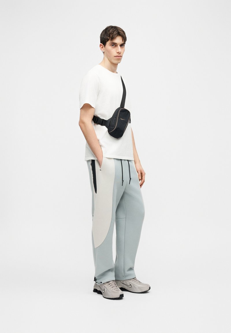 Homme debout portant un t-shirt blanc, un jogging bleu clair et blanc, des baskets beige, et un sac bandoulière noir.