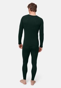 Muž stojící bosý, oblečený v přiléhavém tmavě zeleném dlouhém tričku a odpovídajících legínách, zobrazen zezadu na bílém pozadí.