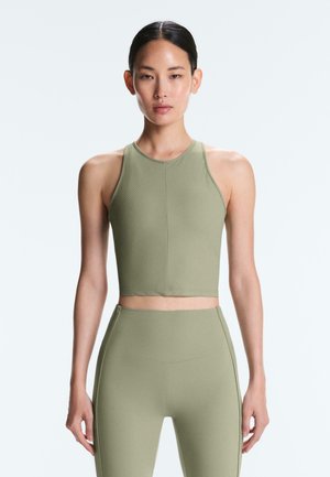 Kobieta o ciemnych włosach, ubrana w oliwkowo-zielony krótki top z wysokim kołnierzem i bez rękawów oraz dopasowane legginsy z wysokim stanem, stojąca na jednolitym białym tle.