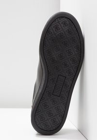 Semelle de chaussure noire avec motif en losange comportant de petits logos et une empreinte de marque rectangulaire "GUESS" au centre.
