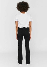 Femme aux cheveux bouclés, vue de dos, portant une chemise blanche à manches courtes, un pantalon noir évasé taille haute et des chaussures blanches.