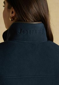 Žena s tmavohnedými vlasmi, oblečená v zlatých kruhových náušniciach a tmavomodrej fleecovej bunde s vystuženým golierom, na ktorom je vzor "JOULES".