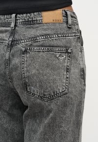 Κοντινή λήψη τσεπής από γκρι τζιν acid-wash με μικρό λογότυπο σε σχήμα καρδιάς και δερμάτινο ταμπελάκι με την επιγραφή «ROXY» στη ζώνη μέσης.