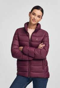 Bordeauxfärgad puffjacka med hög krage, dragkedja och elastiska ärmslut. Utrustad med horisontell quiltad design och logotypbroderi på bröstet.